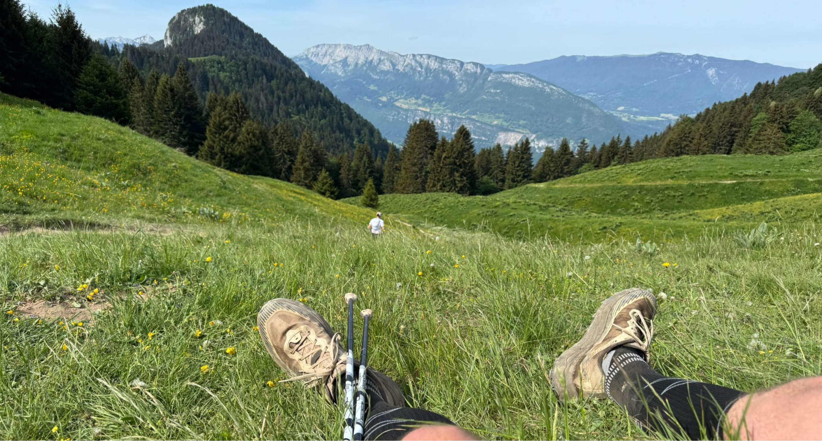 Une personne se repose dans l’herbe avec ses chaussures et bâtons face au paysage montagneux.