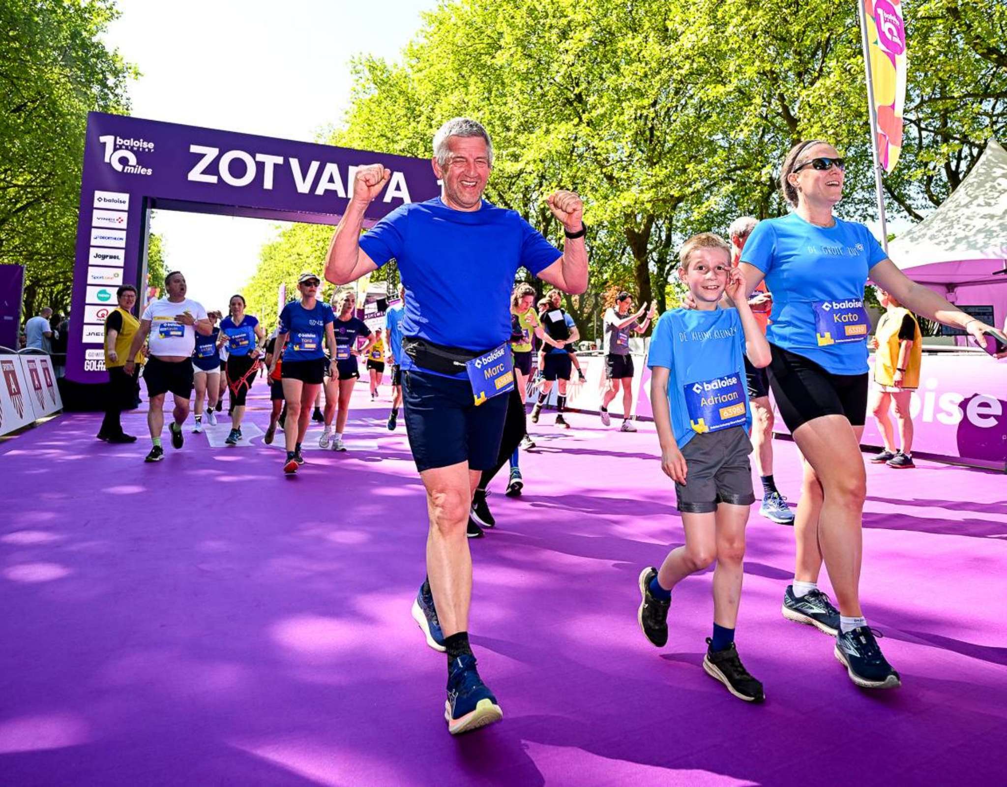 Deelnemers steken juichend de finishlijn over onder de paarse boog van “ZOT VAN A 10 miles”.