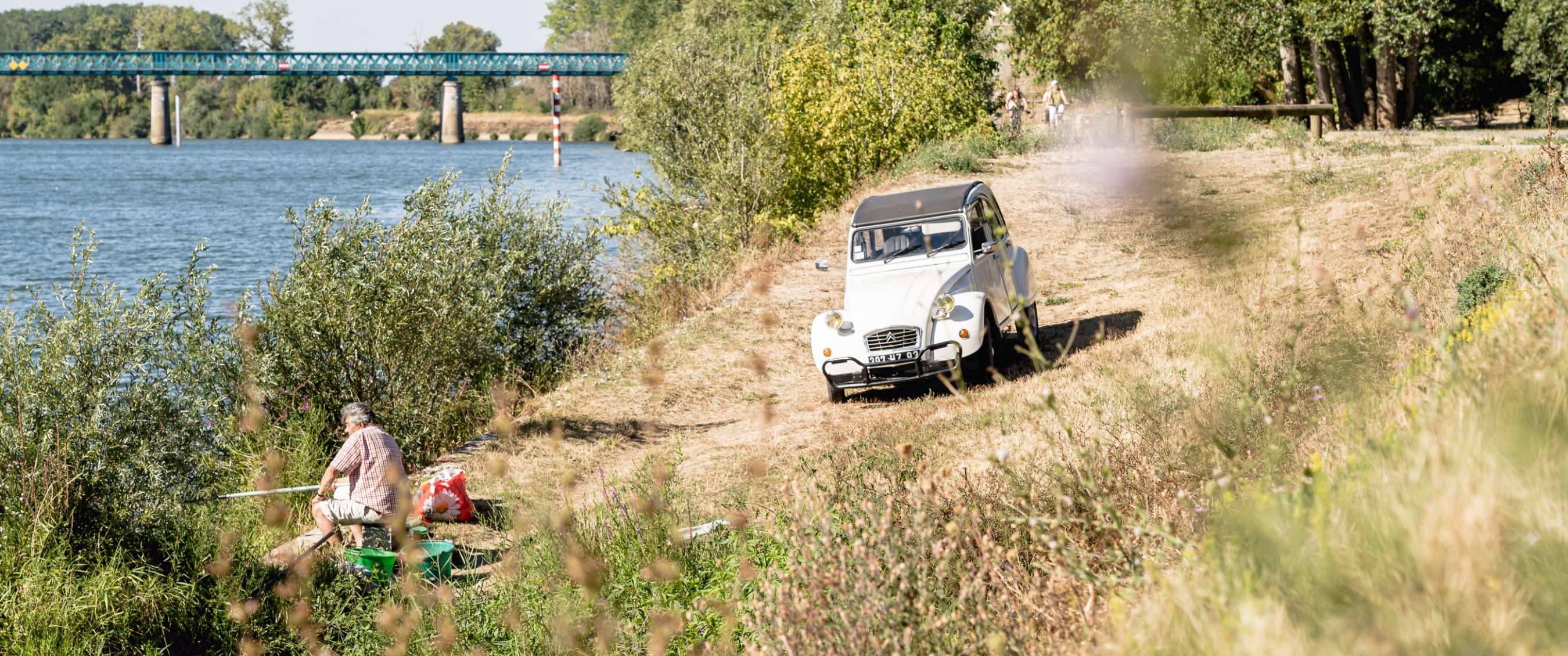 Witte Citroën 2CV op een oeverpad langs de rivier, met een visser op de voorgrond.