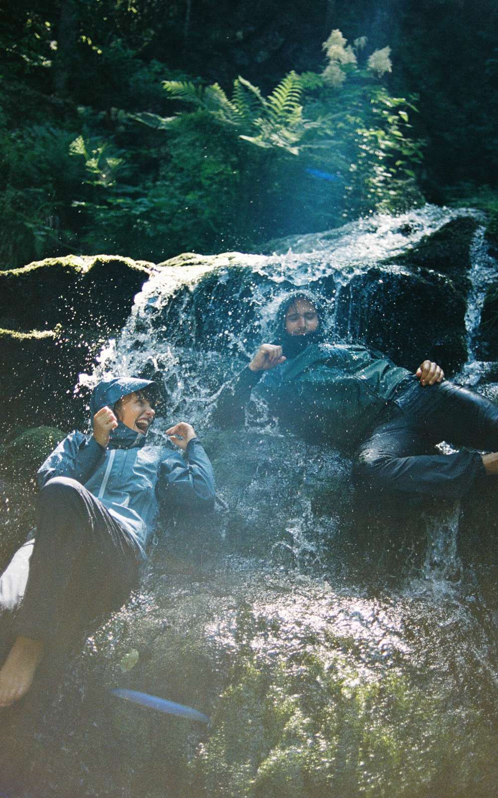 Deux personnes en vêtements imperméables sont assises sous une petite cascade entre des rochers sombres.