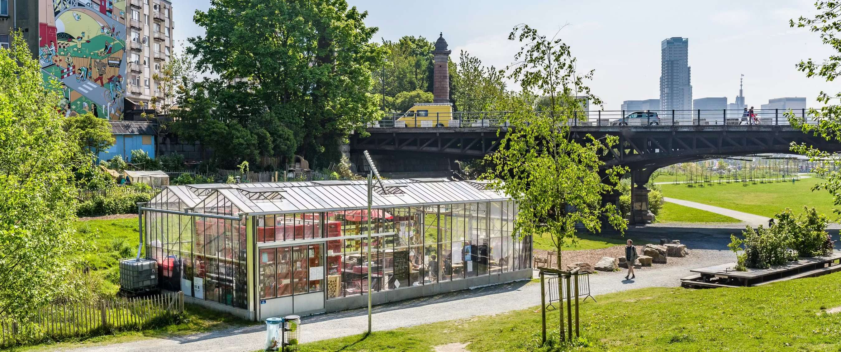 Serre en verre dans un parc urbain sous un pont avec bâtiments en arrière-plan à Bruxelles.