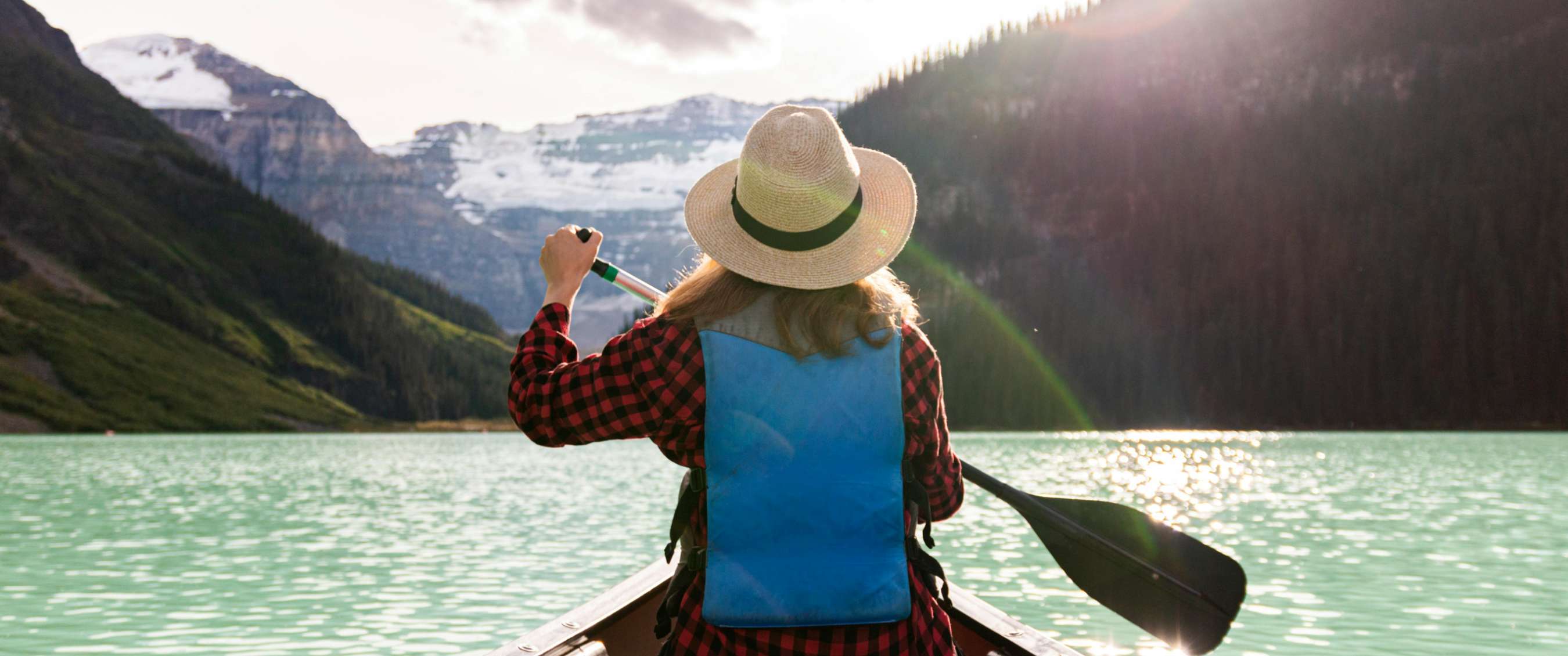 Une femme avec un chapeau et une chemise à carreaux pagaie sur un lac de montagne turquoise en direction de montagnes enneigées.
