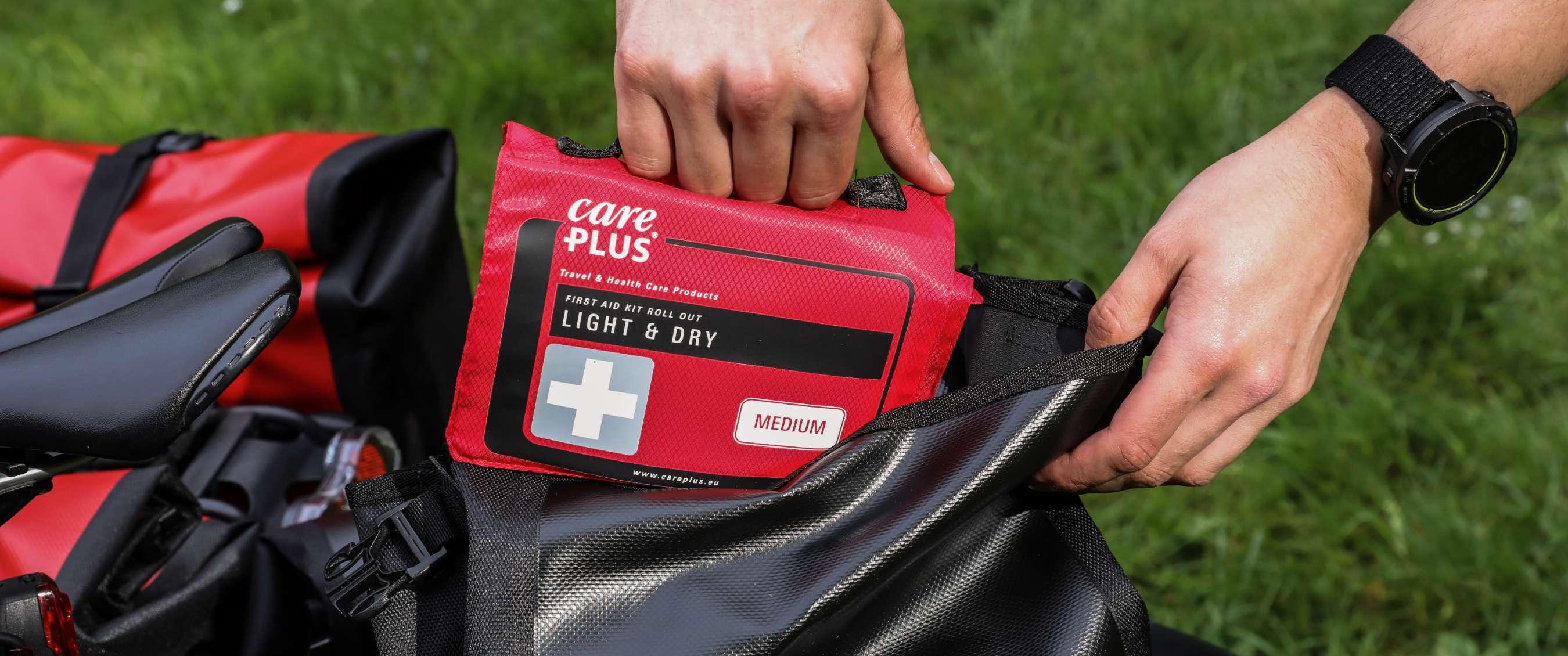 Hand plaatst Care Plus EHBO-set Light & Dry Medium in zwarte fietstas op gras