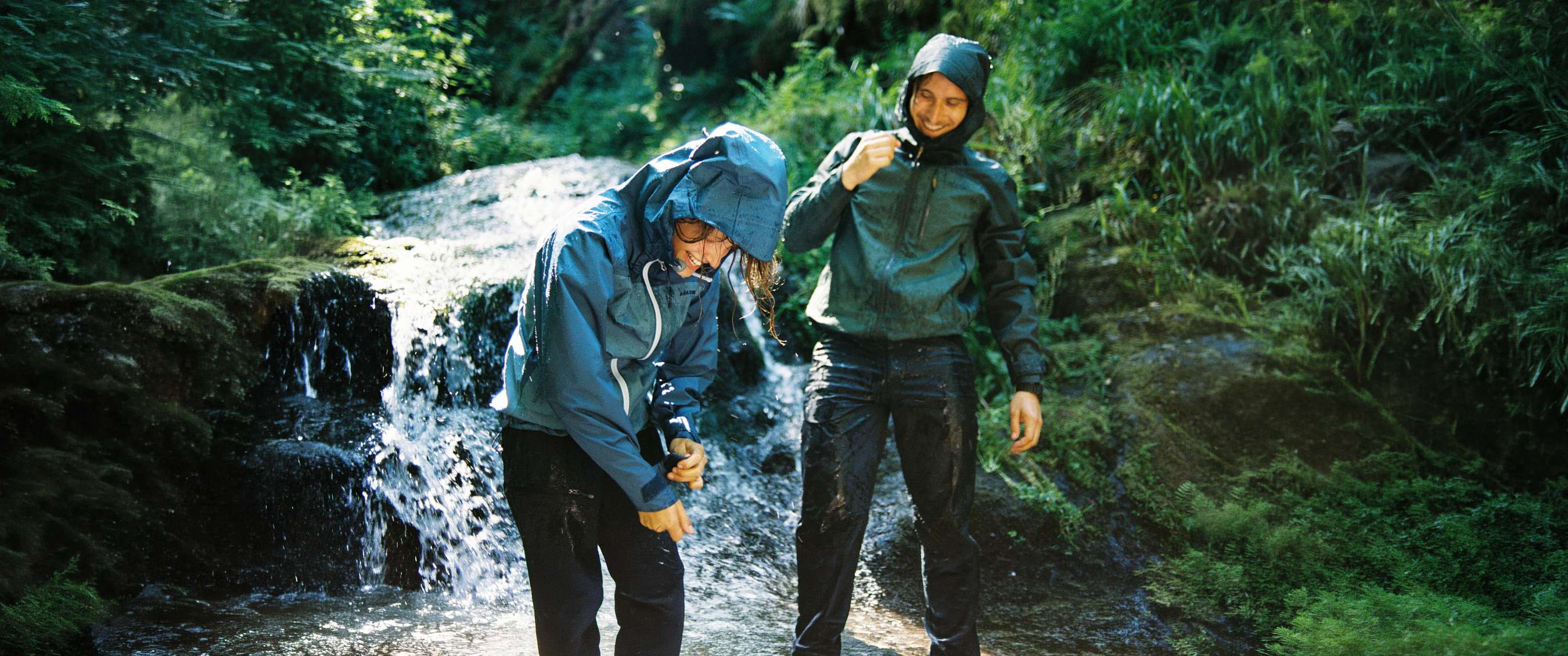 Deux personnes en vêtements imperméables se tiennent près d’une cascade entourée de verdure.