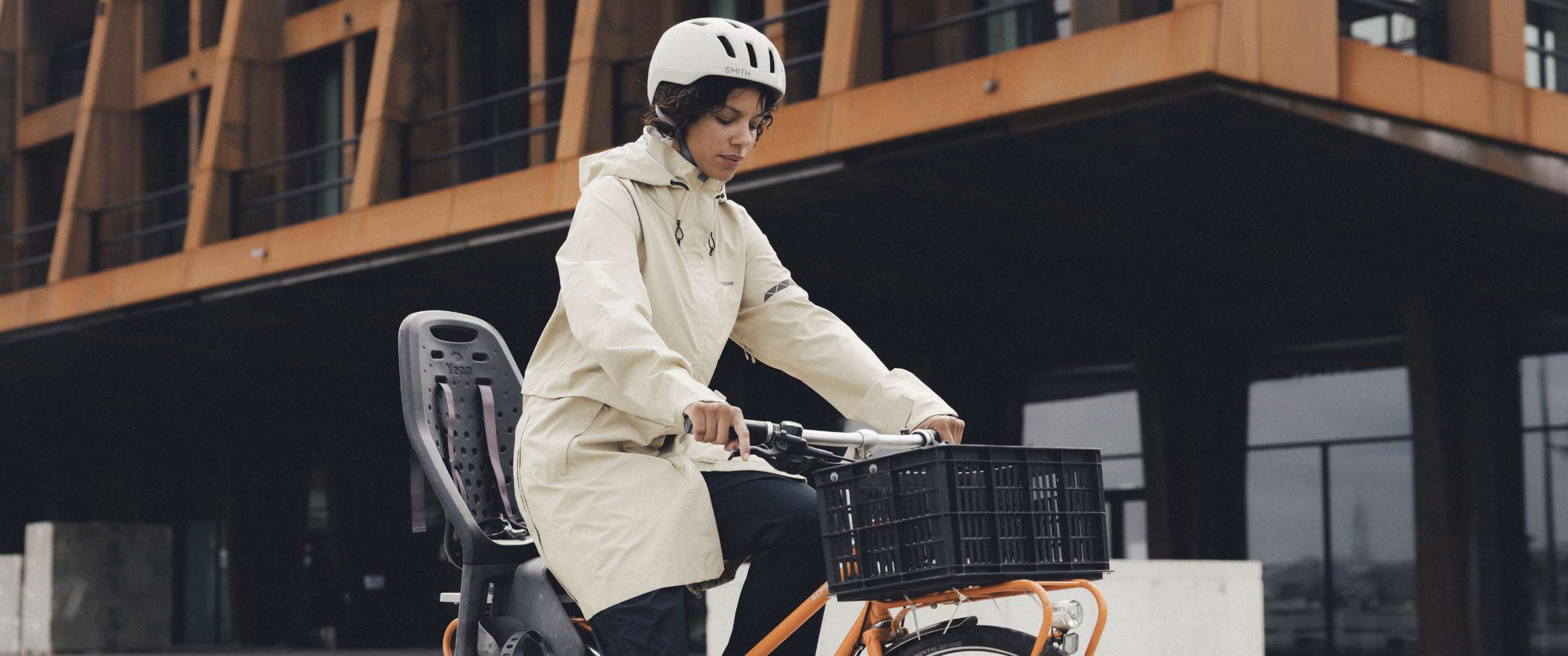 Vrouw fietst door de stad met regenjas en helm op, voor een modern gebouw.