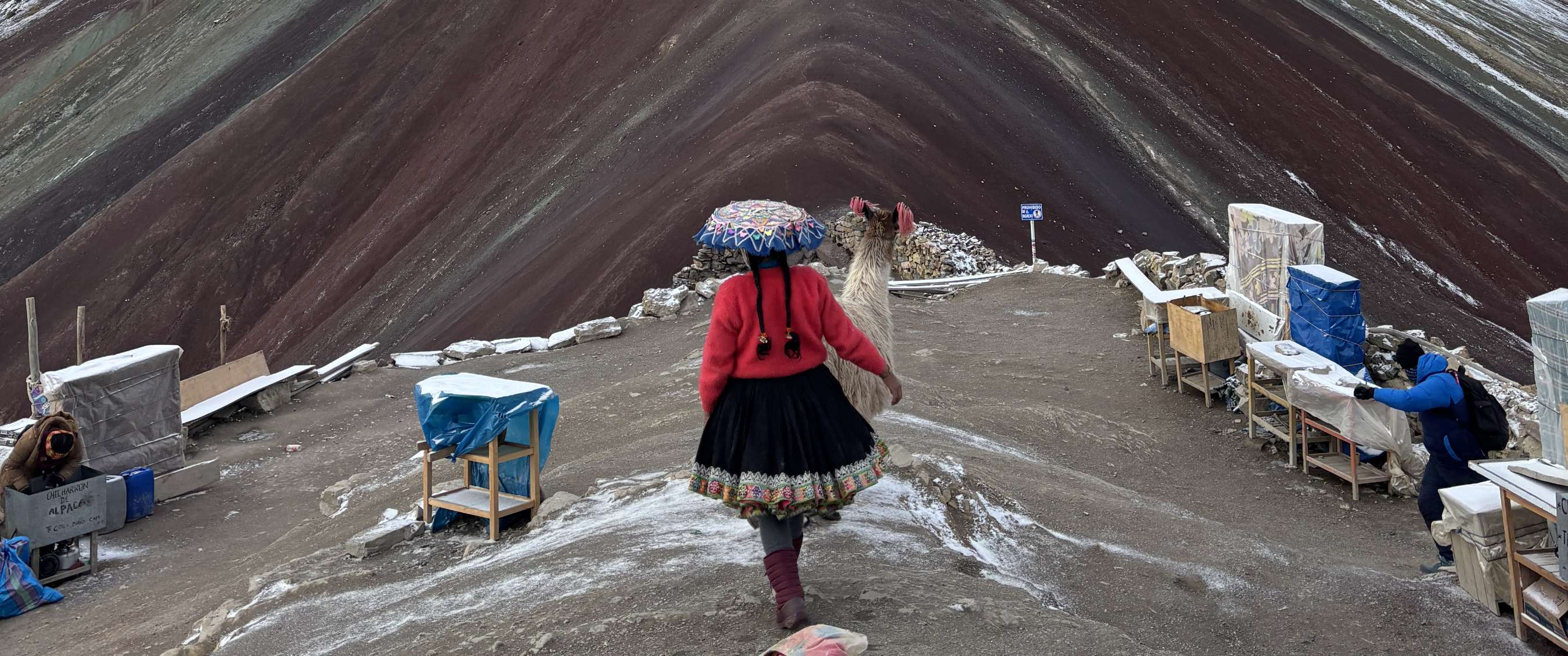 Persoon in traditionele kleurrijke kleding met lama op bergpad bij gestreepte helling.