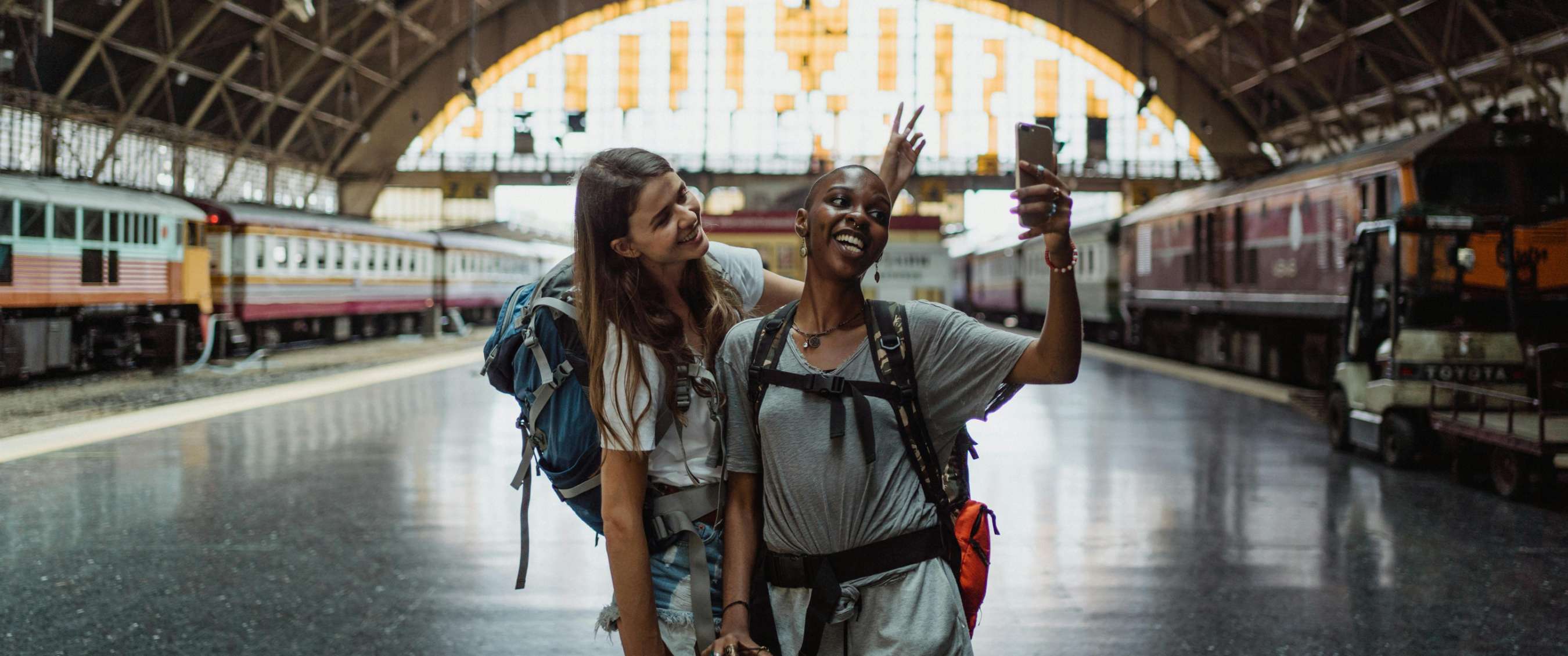 Twee beelden: twee reizigers met rugzakken maken een selfie op een treinstation tussen stilstaande treinen; grote stationshal met perrons en treinen op de achtergrond.