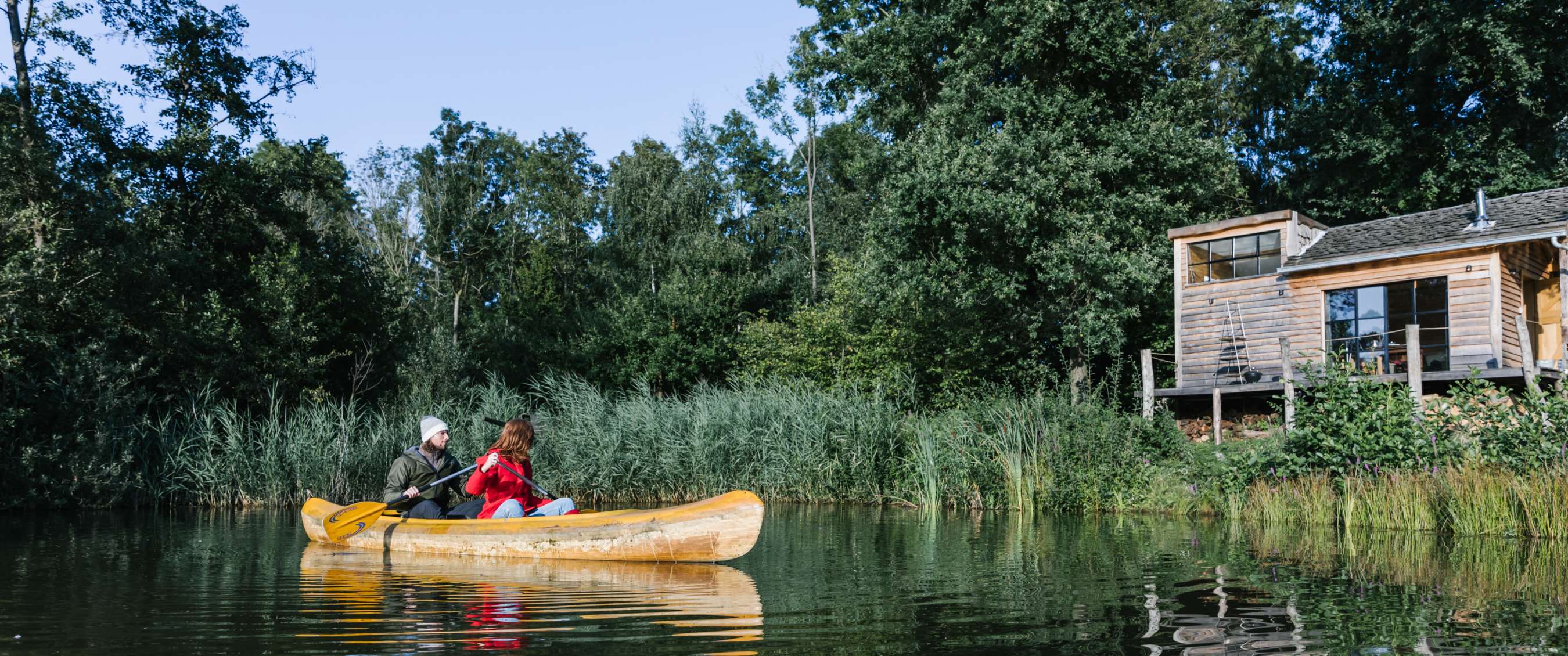 Canoë avec deux personnes sur un étang dans un environnement verdoyant, avec un chalet au bord de l’eau.