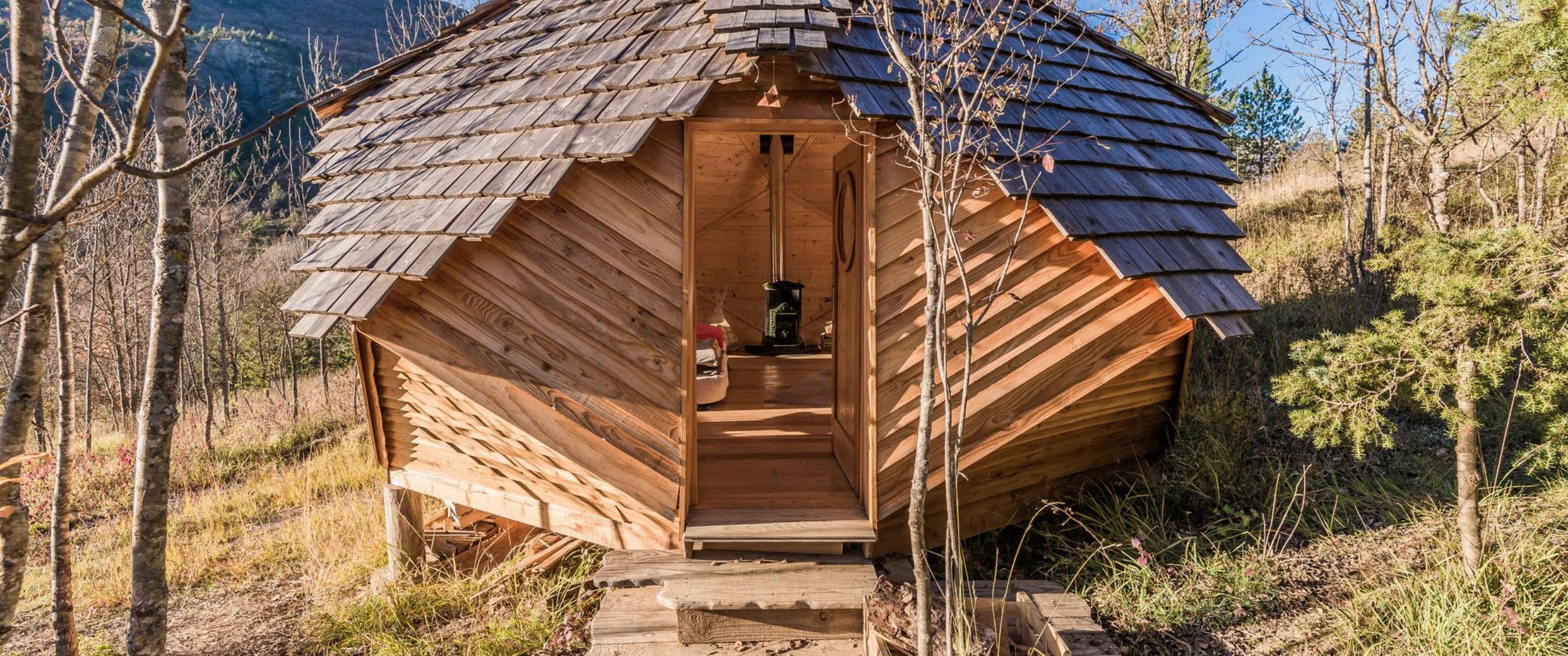 Cabane circulaire en bois avec toit en bardeaux dans un environnement naturel montagneux.