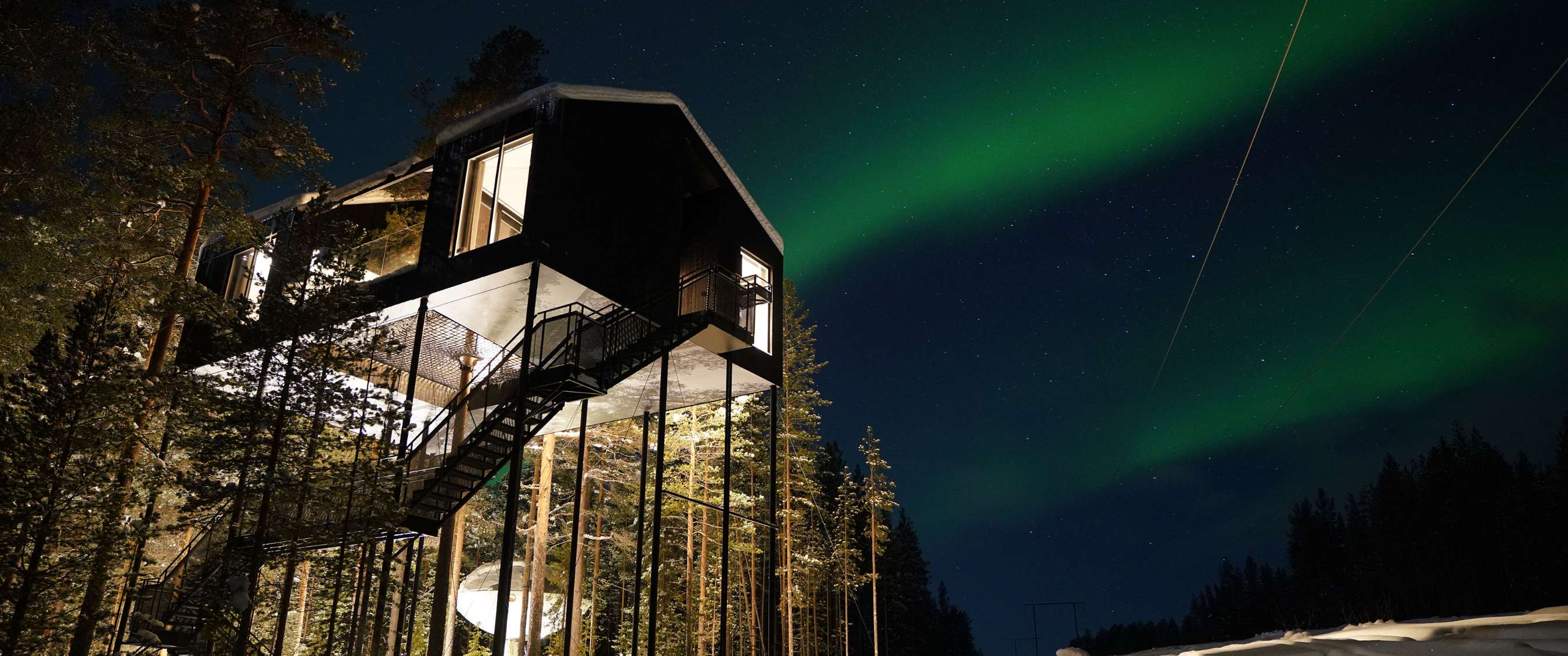 Cabane perchée en bois dans une forêt enneigée, intérieur éclairé et aurore boréale en arrière-plan.