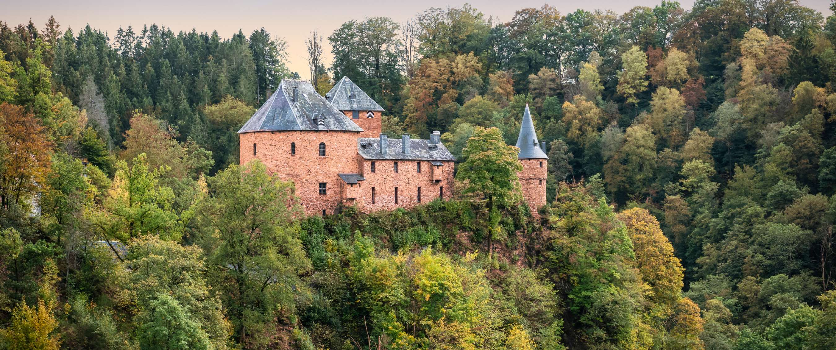 Rood stenen kasteel met torens staat op een beboste heuvel tussen herfstkleurige bomen.