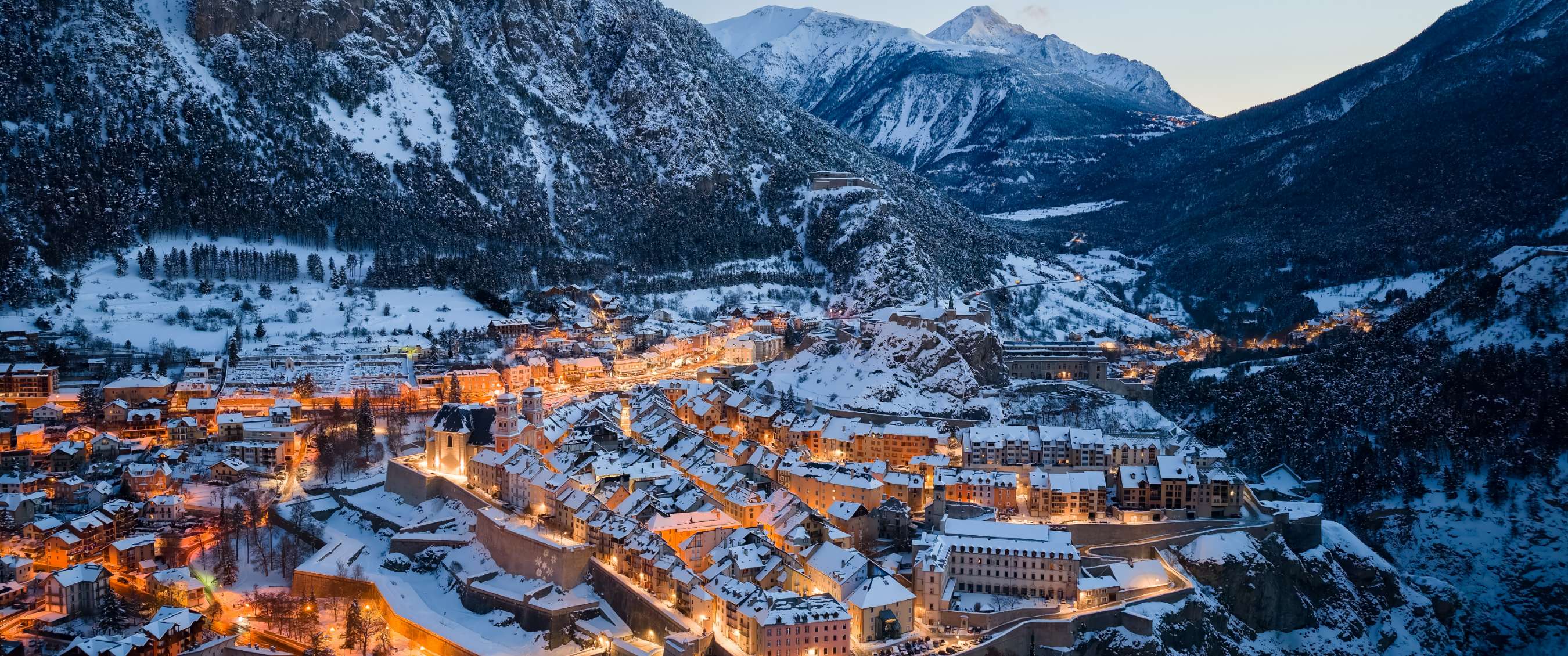Besneeuwde stad met verlichte straten tussen beboste bergen bij schemering.