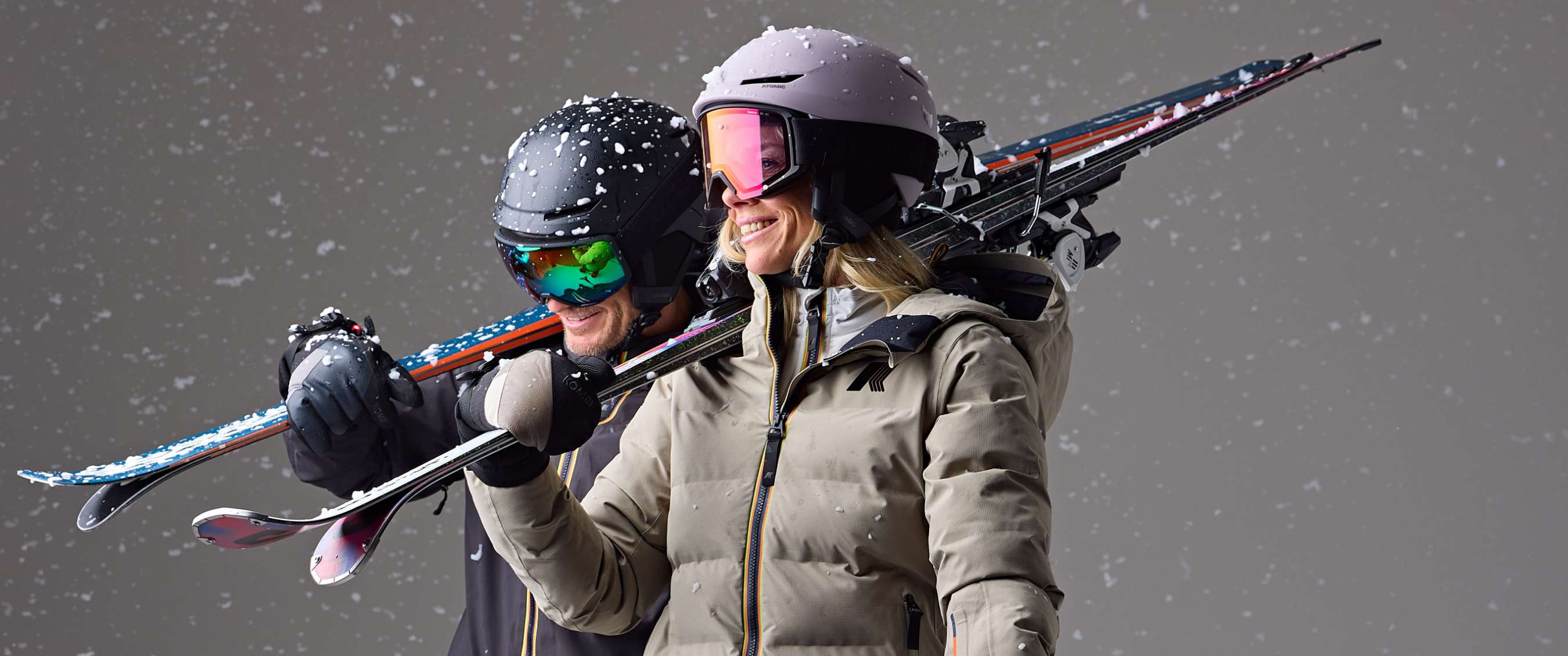 Deux skieurs portent leurs skis sur les épaules avec des flocons de neige autour d’eux.