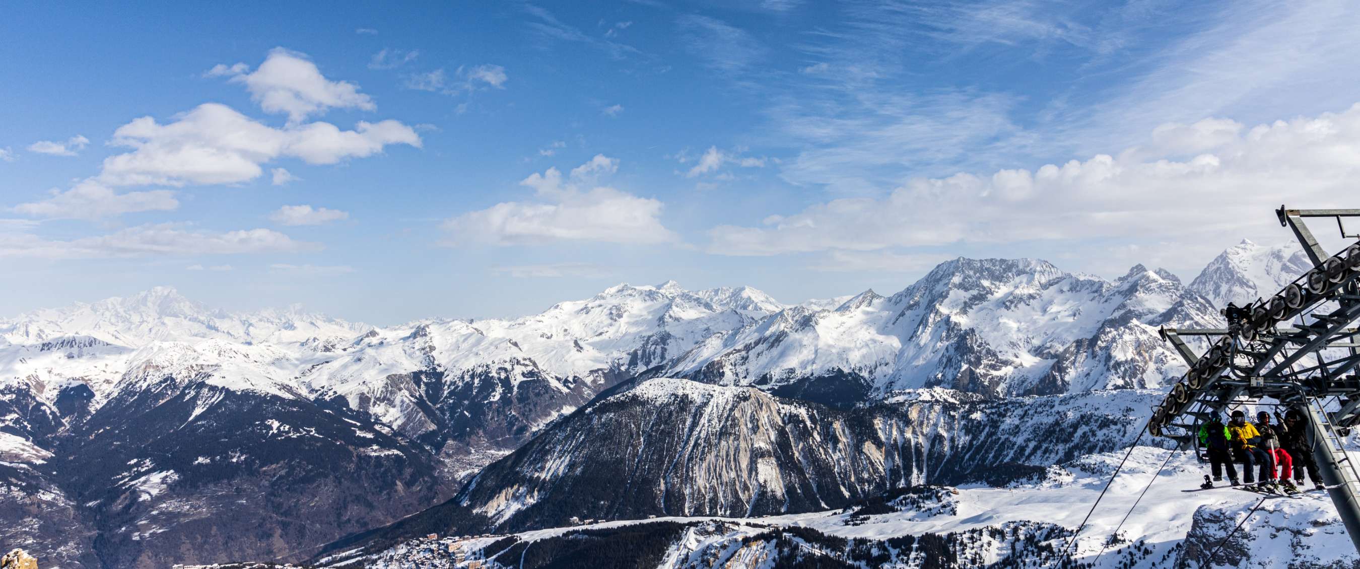 Un télésiège avec des passagers au premier plan, des montagnes enneigées en arrière-plan et un ciel bleu clair avec des nuages blancs aux Trois Vallées.