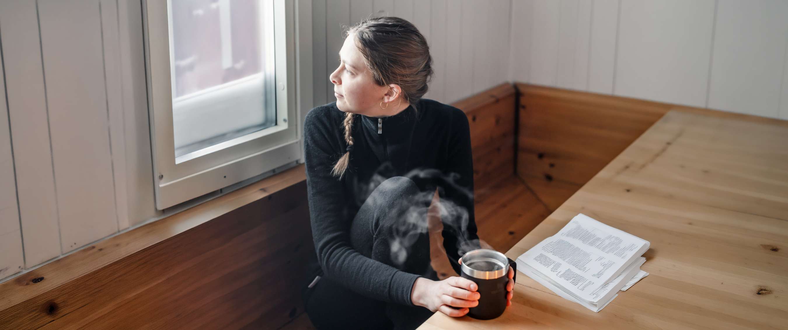 Personne en vêtement thermique noir assise près d’une fenêtre avec une tasse fumante.